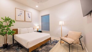 Une chambre à coucher comprenant un lit, une chaise, une table de chevet, une lampe, une fenêtre avec des stores et deux tableaux représentant des oiseaux, accrochés au mur.