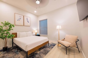 A bedroom with a bed, a chair, a nightstand, a lamp, a window with blinds, and two wall-mounted bird paintings.