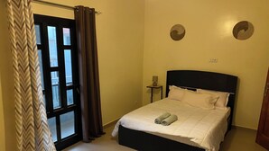 Ein ordentlich bezogenes Bett mit weißen Leinentüchern und zwei aufgerollten Handtüchern, ein Nachttisch mit Lampe und eine Tür mit Vorhängen.