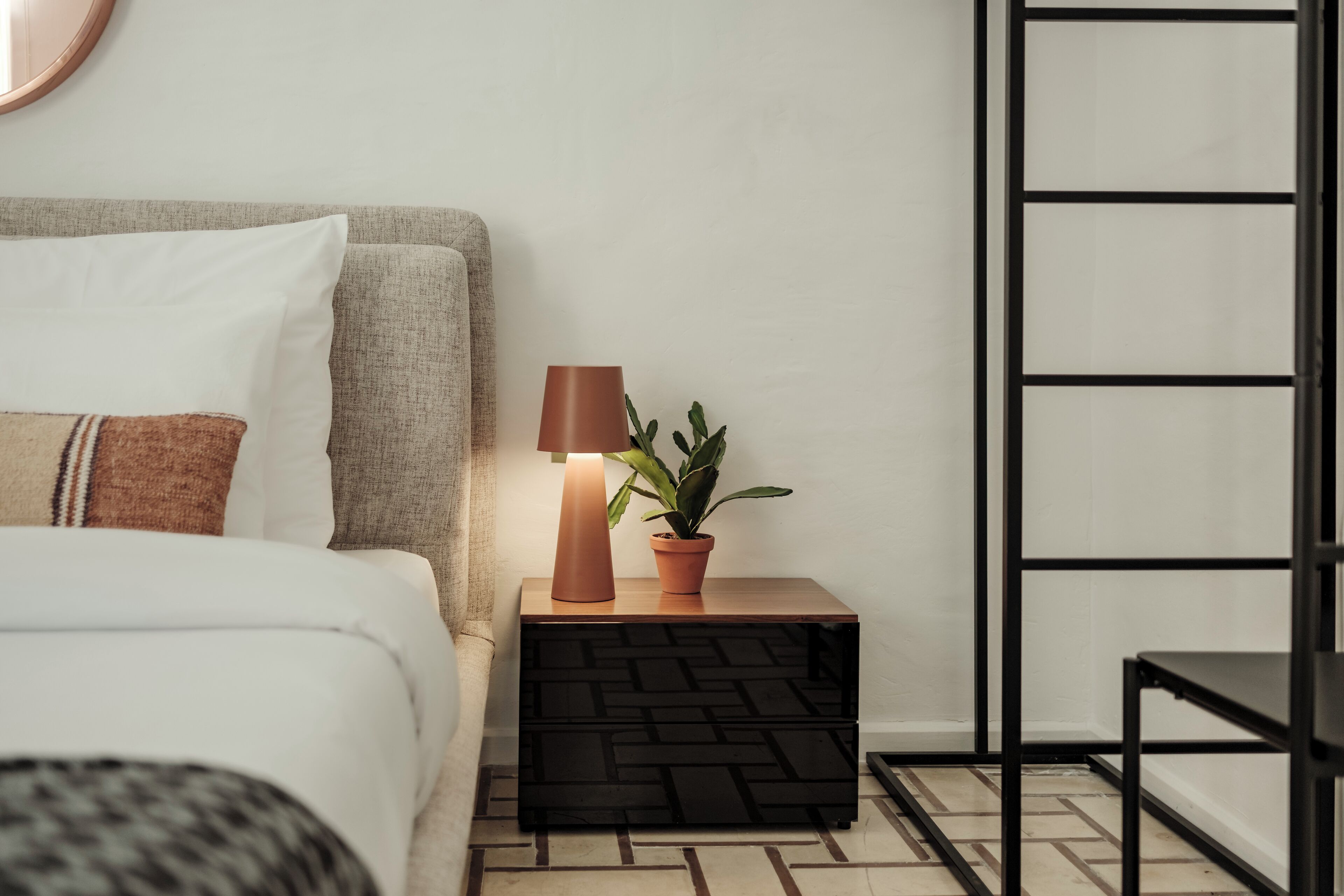 Un dormitorio ordenado con cama, mesita de noche, lámpara y planta en maceta.