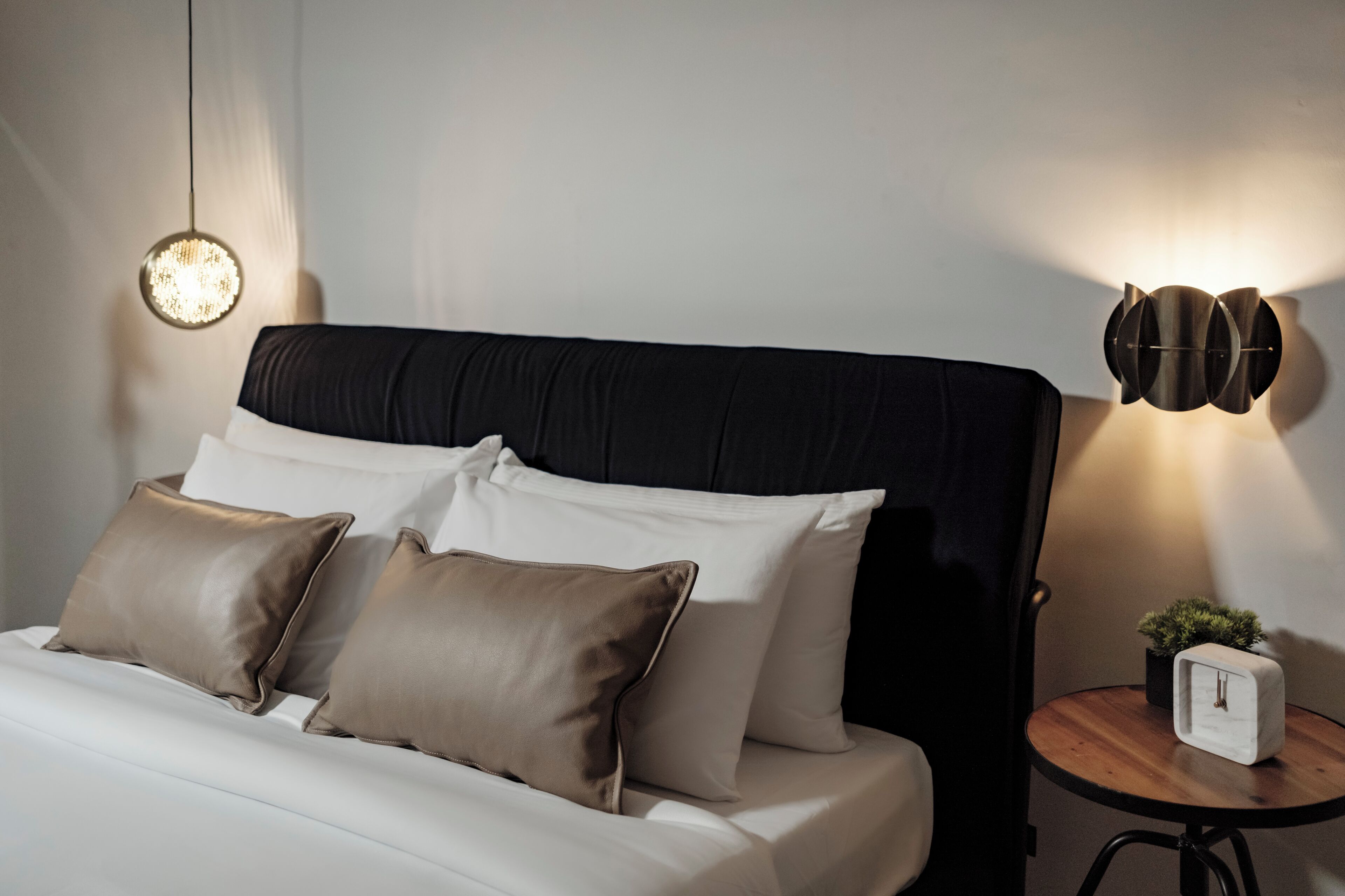 A neatly made bed with white and brown pillows, a round table with a plant, and a wall-mounted light fixture.