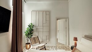A hotel room with a bed, a potted plant, a chair, a wall-mounted TV, and a door. - Ellul Living (Floriana)