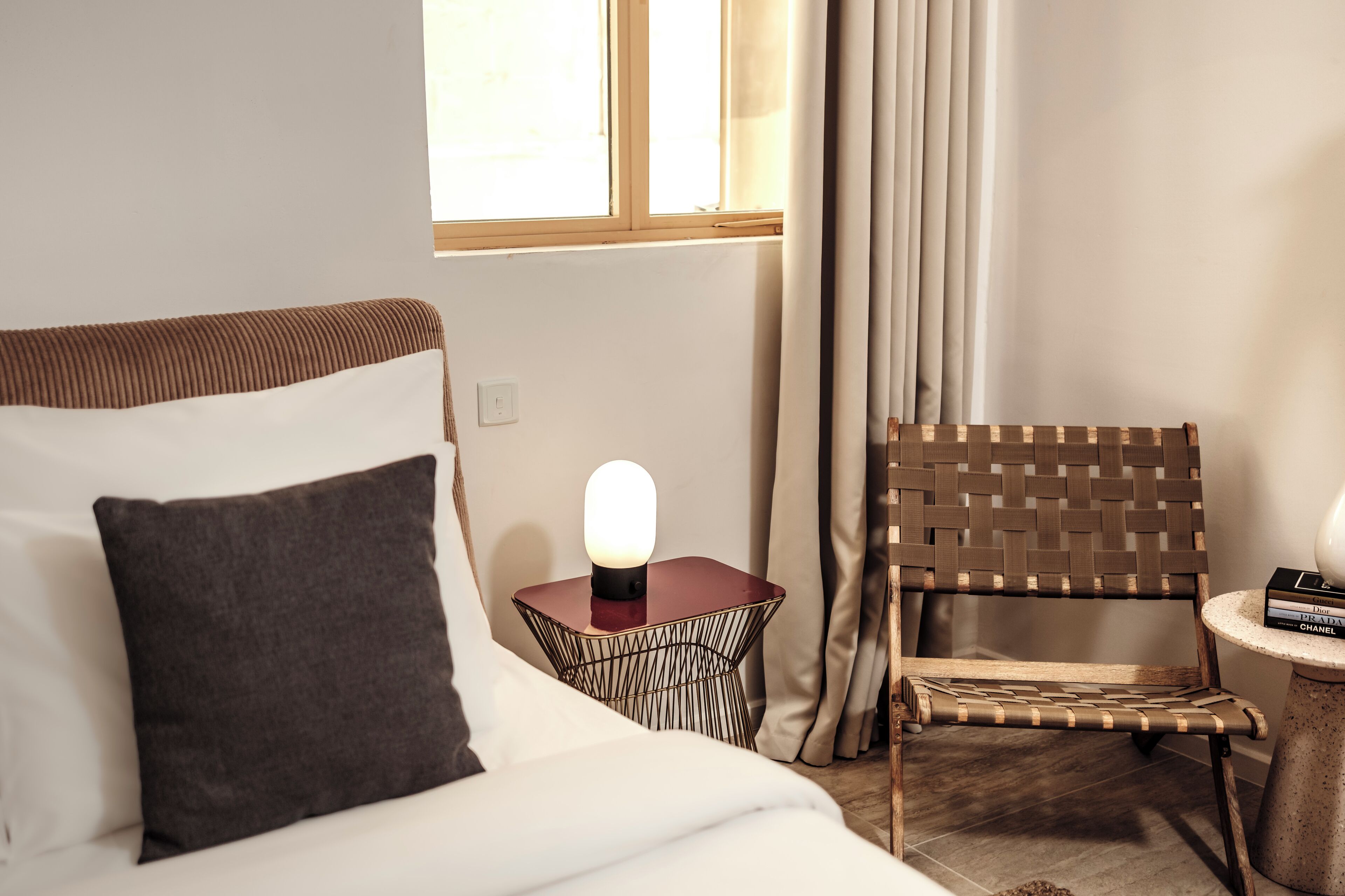 A hotel room with a bed, a side table with a lamp, a wicker chair, and a small round table with books.