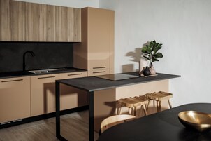 Una cocina moderna con encimera negra, armarios de madera y grifo negro. Hay una pequeña mesa con taburetes y una planta en maceta.