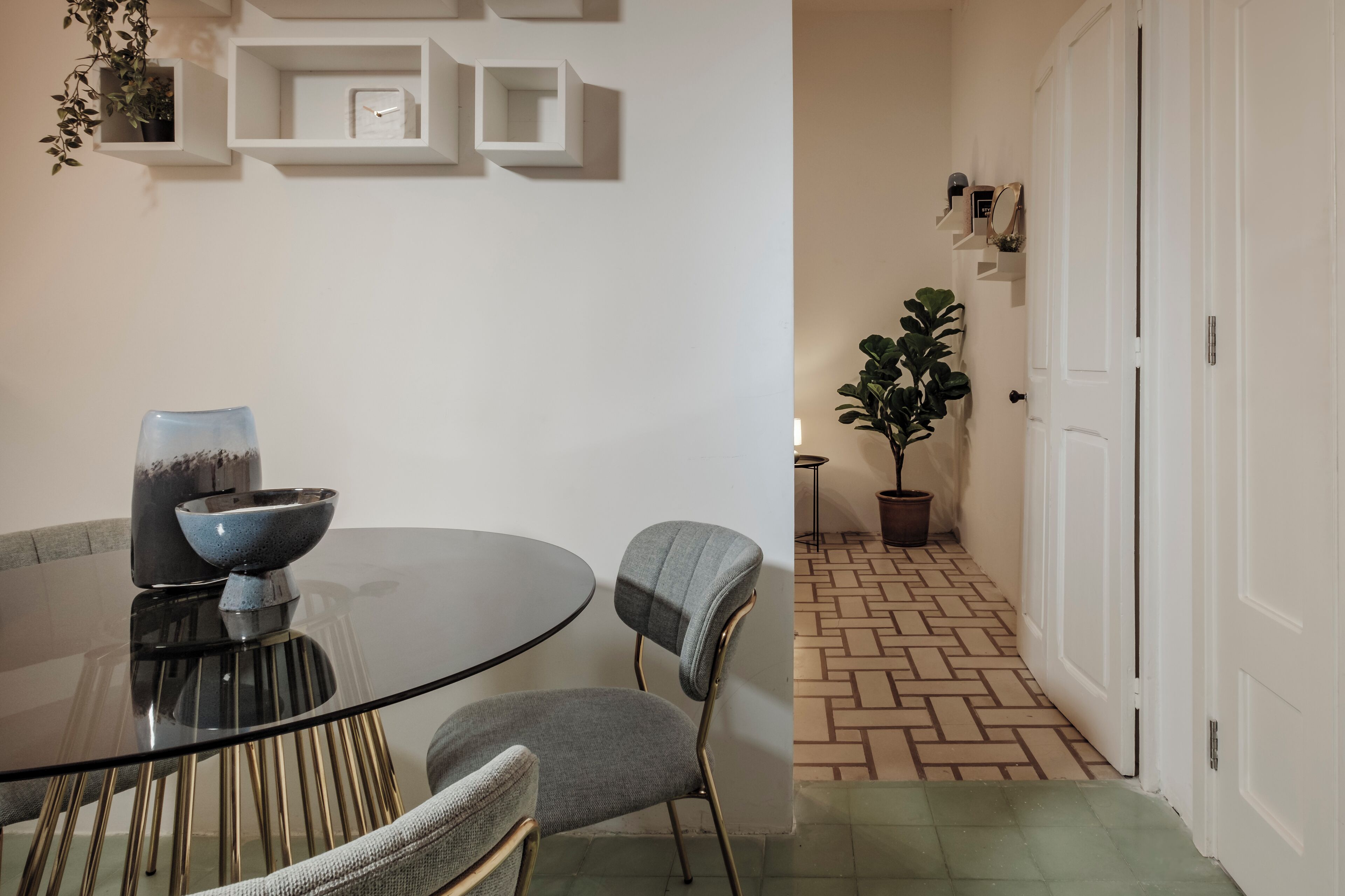 A dining area with a round glass table, two chairs, and a vase. There are white walls with shelves and a potted plant in the background.