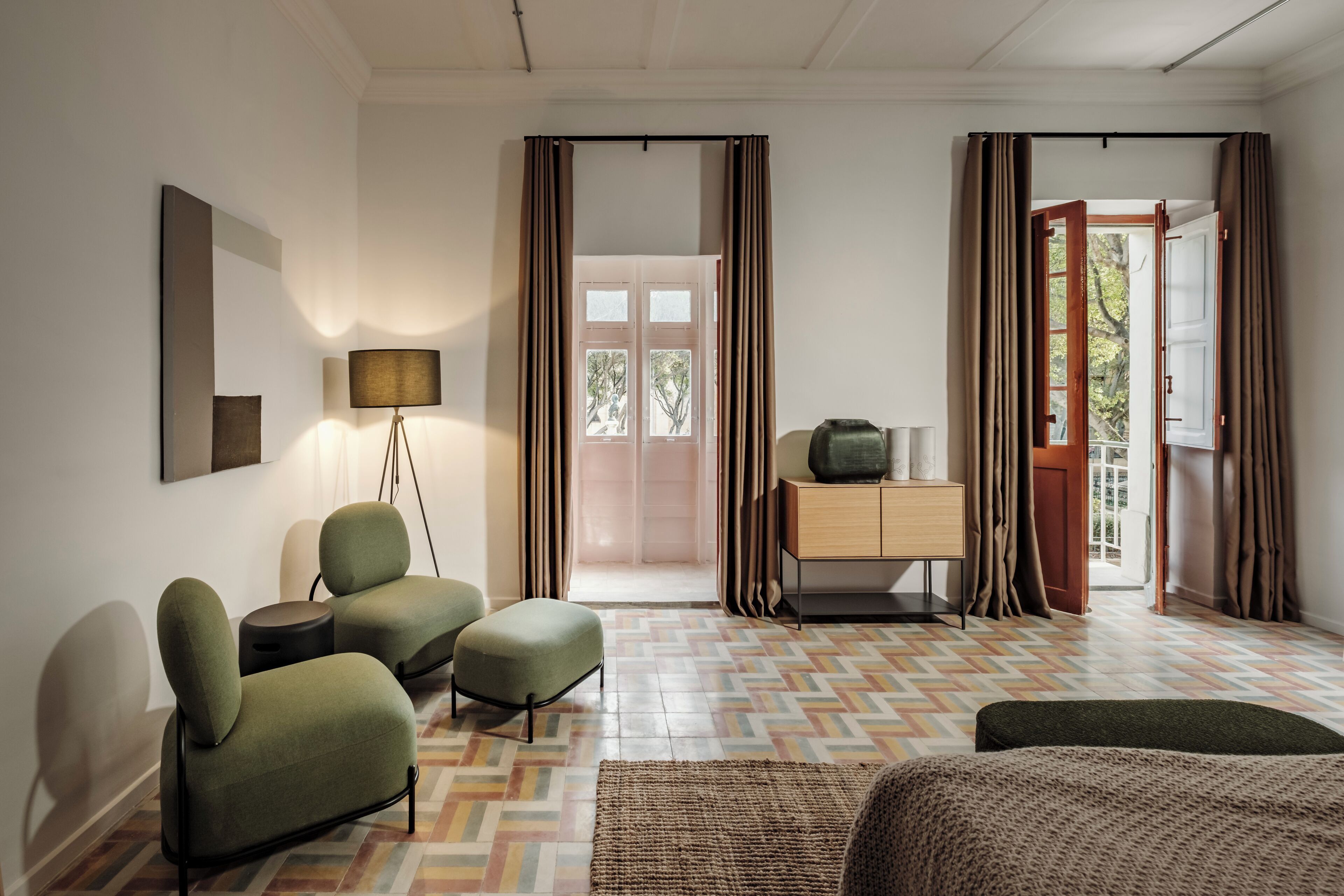 Habitación de hotel moderna con suelo de patrón espiga, una cama, dos sillones, una mesita auxiliar, una lámpara, un aparador y vistas a través de un vano.