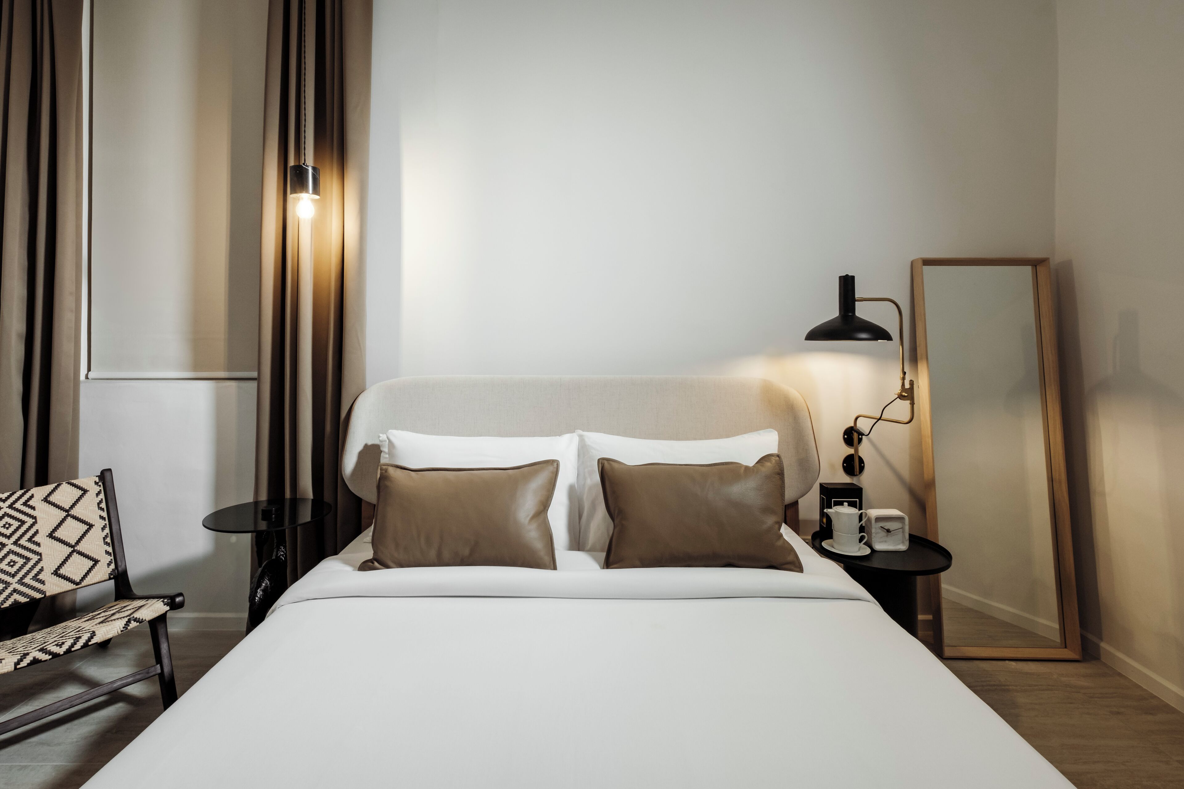 A neatly arranged hotel room with a bed, a side table with a lamp, a chair, a mirror, and a window with curtains.
