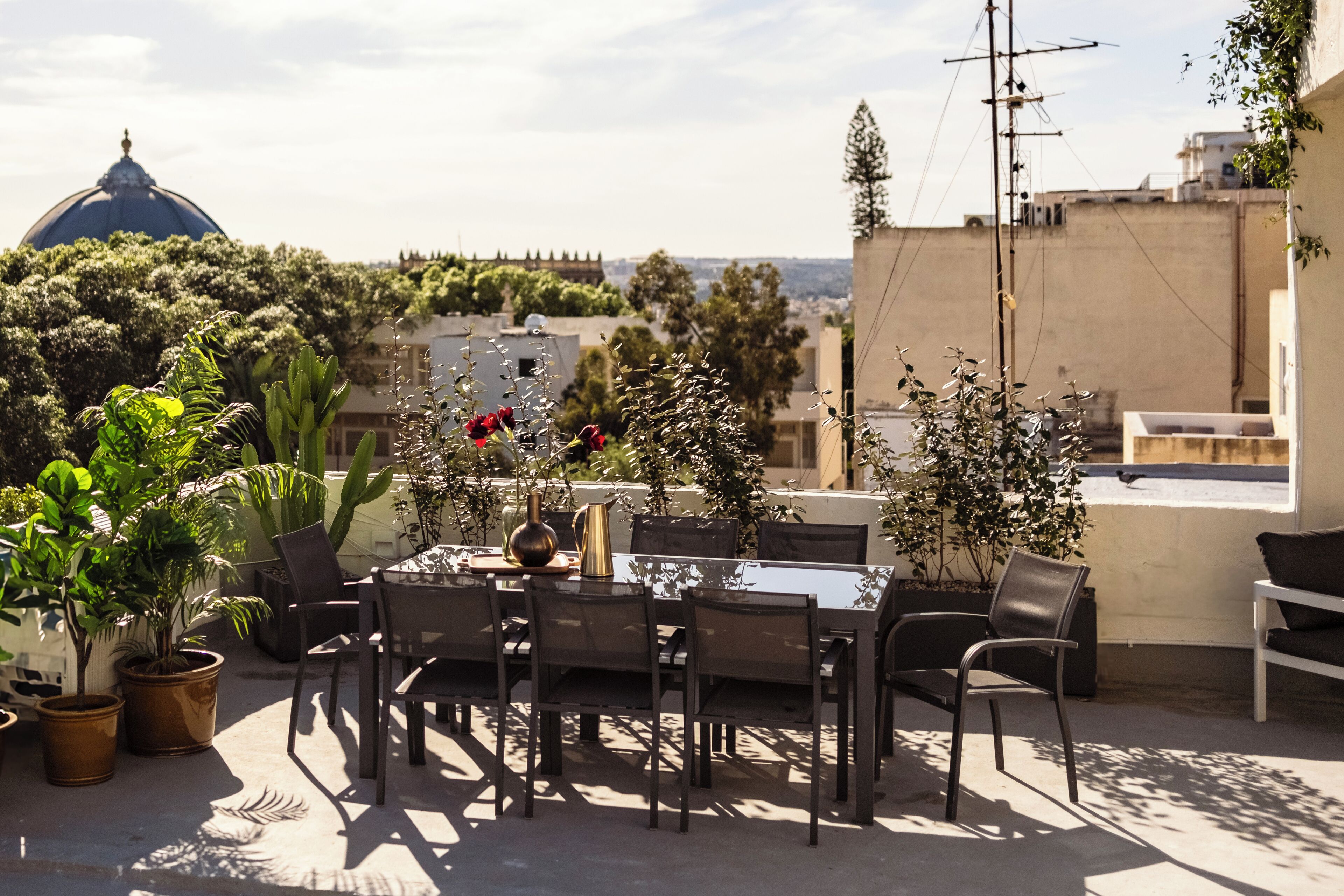 Um pátio externo com mesa e cadeiras, rodeado por plantas em vasos e vista para a cidade.