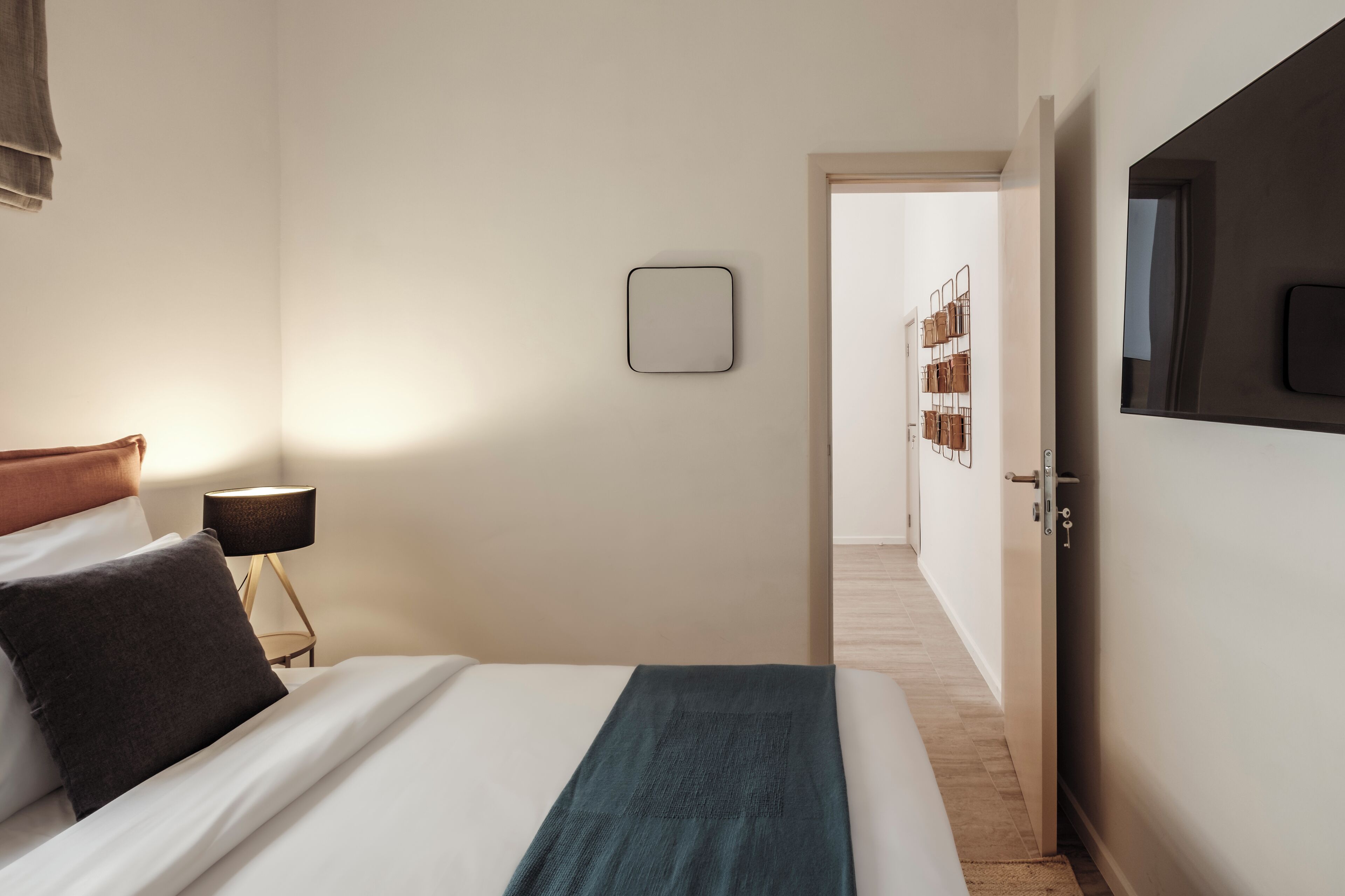 Ein ordentlich bezogenes Bett mit weißer und türkisgrüner Decke, einer Nachttischlampe und einem an der Wand montierten Fernseher.