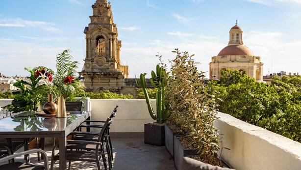 A rooftop terrace with a dining table, chairs, and potted plants. In the background, there are two prominent buildings with domes and spires. - Ellul Living (Floriana)