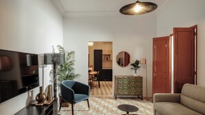 A modern living room with a flat-screen TV, a blue armchair, a sofa, a patterned rug, and a dining area in the background. - Ellul Living (Floriana)