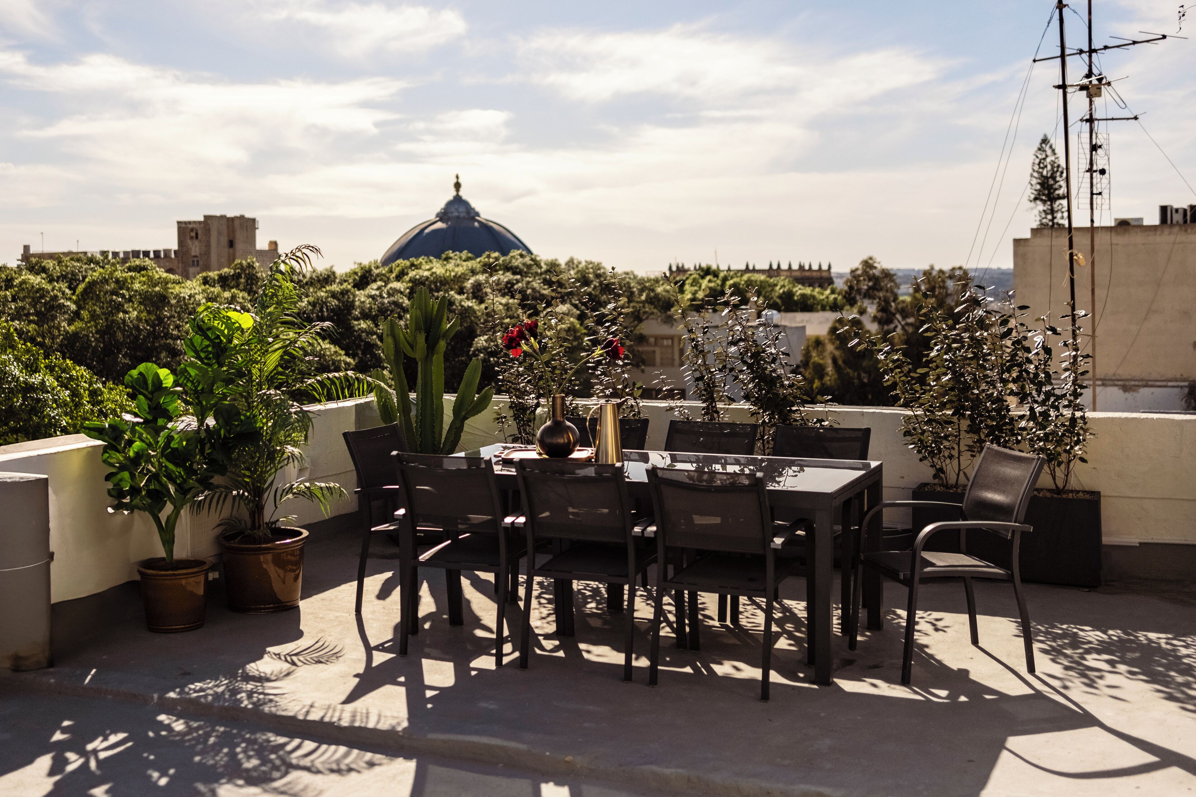 Terraza en la azotea con mesa y sillas, rodeada de macetas con plantas y vistas a una cúpula y edificios a lo lejos.