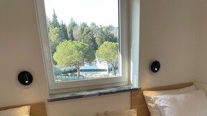 Ein Schlafzimmer mit zwei Betten, einem Fenster mit Blick auf Bäume und ein Gebäude, sowie ein Nachttisch mit einer Blumenvase.