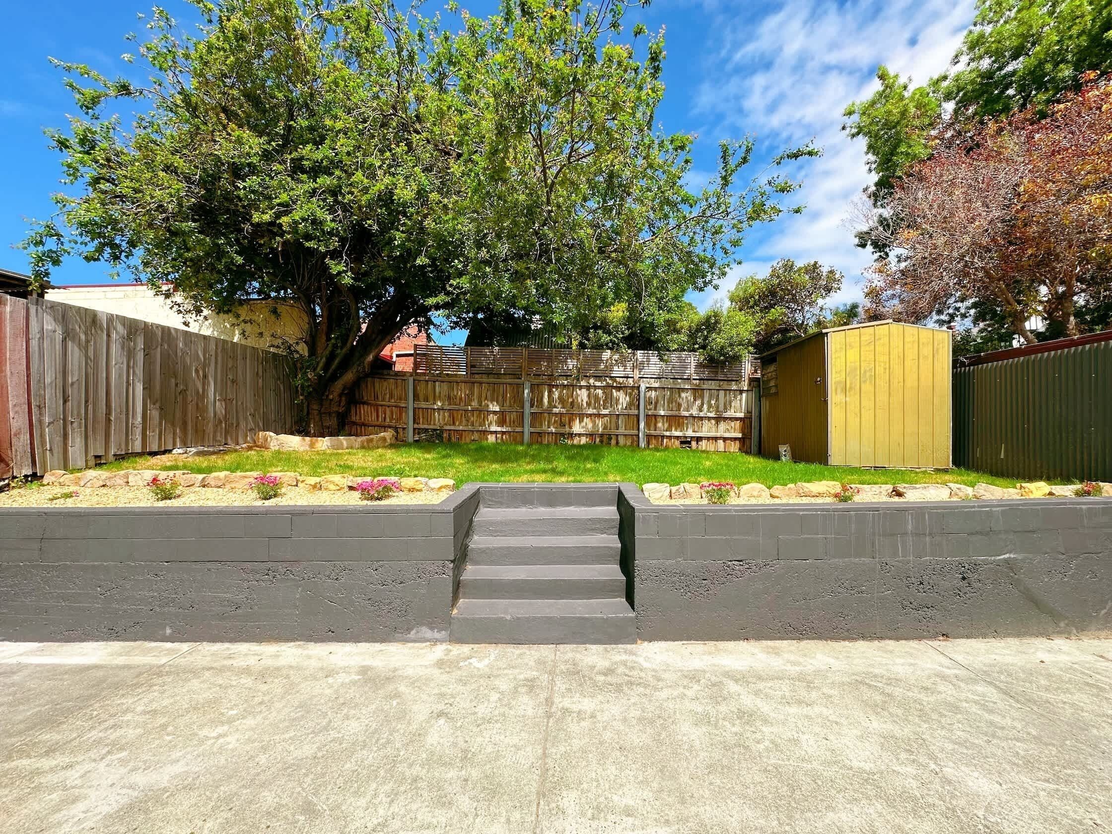A backyard with a wooden fence, a tree, a shed, and a concrete pathway.