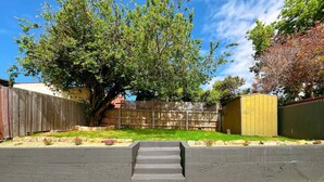Un jardin avec une clôture en bois, un arbre, un abri de jardin et un chemin en béton.