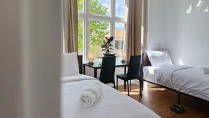 Une chambre d’hôtel avec deux lits, un bureau avec une lampe, une chaise et une vue sur les arbres à travers de grandes fenêtres.