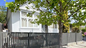 Une maison d’habitation à l’extérieur blanc, avec une clôture en bois et un arbre devant.
