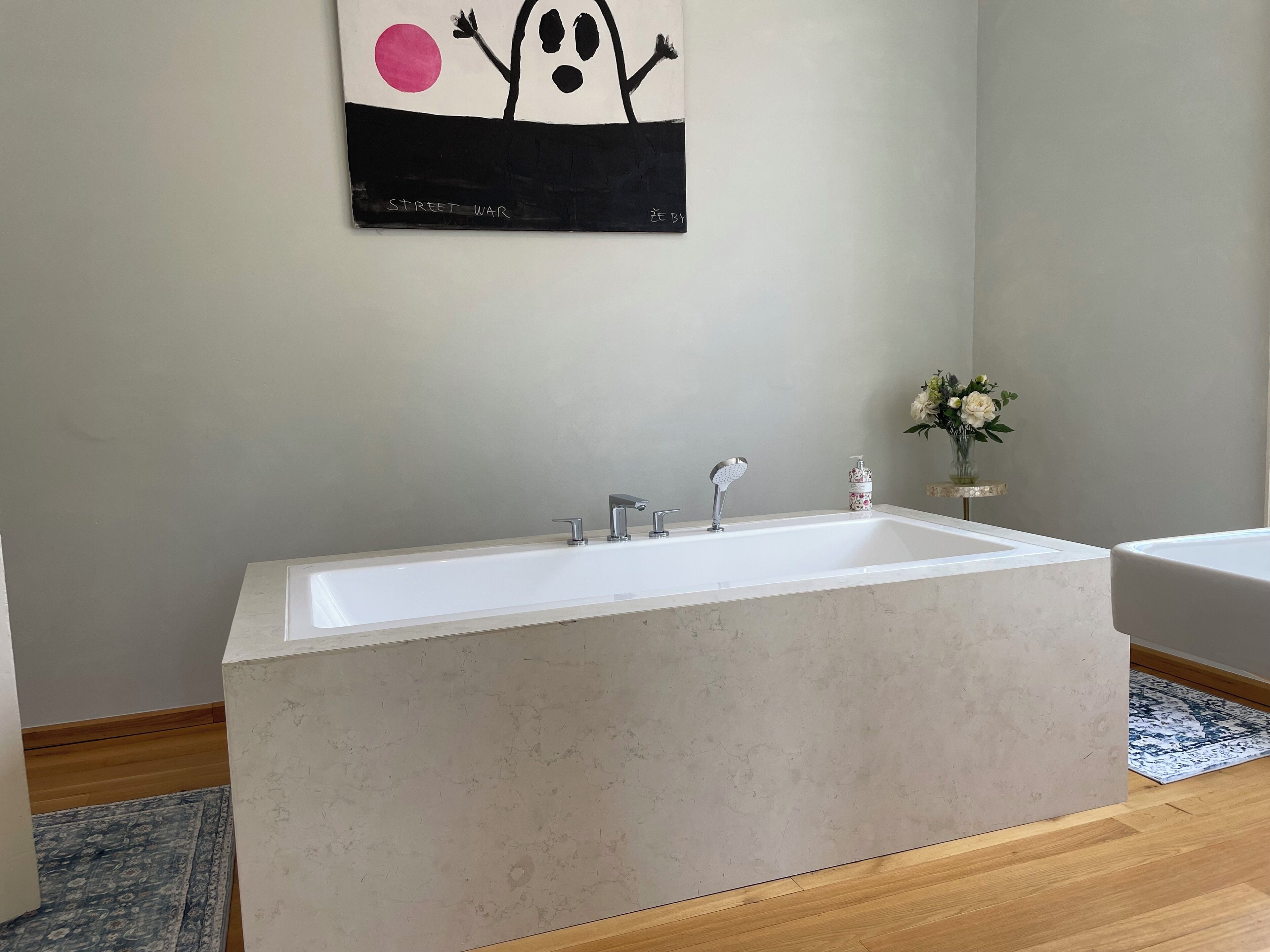 A modern bathroom with a large bathtub, a small sink, and a toilet. There is a painting on the wall and a vase with flowers on a side table.