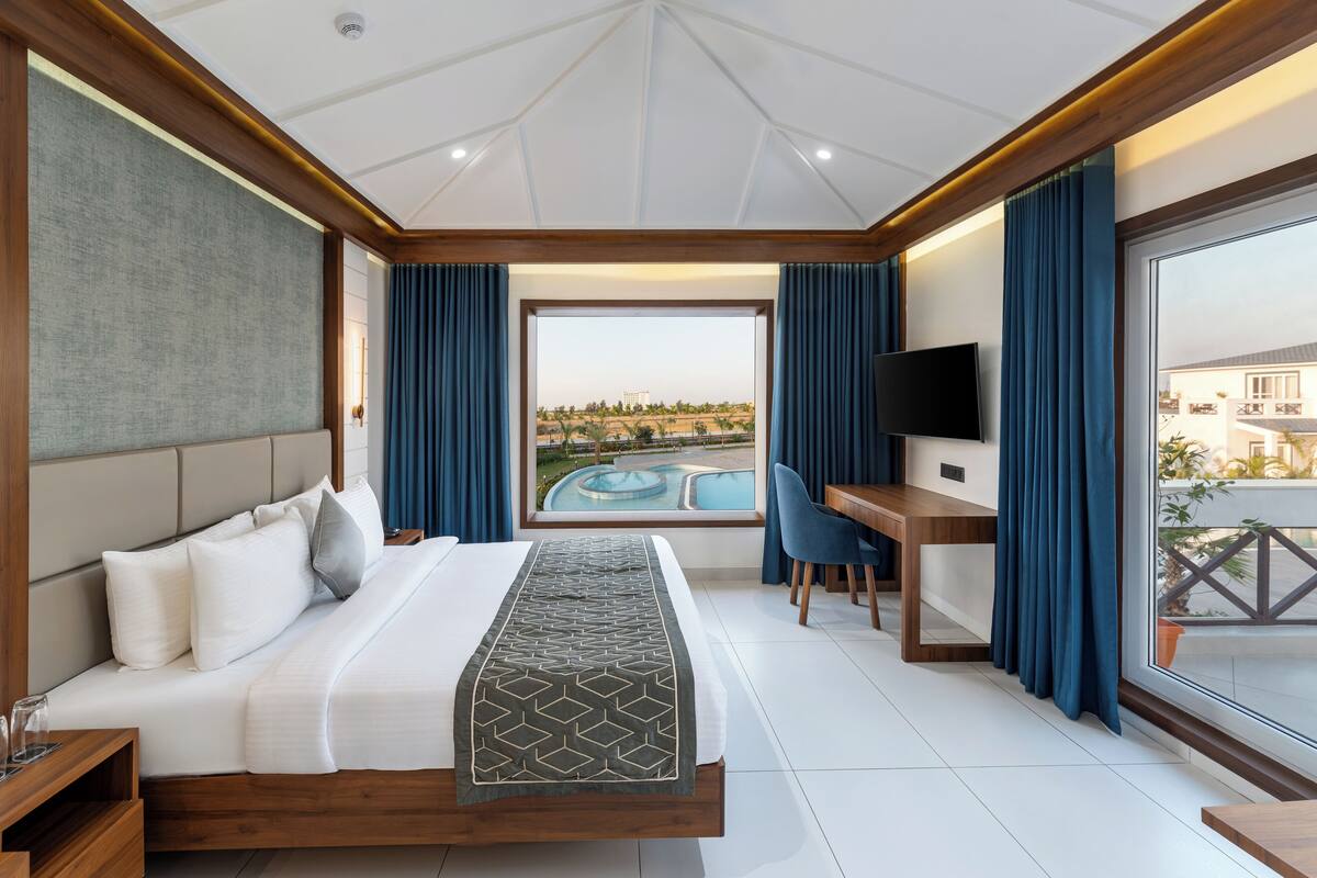 A modern hotel room with a large bed, a desk, a TV, and a view of a swimming pool.
