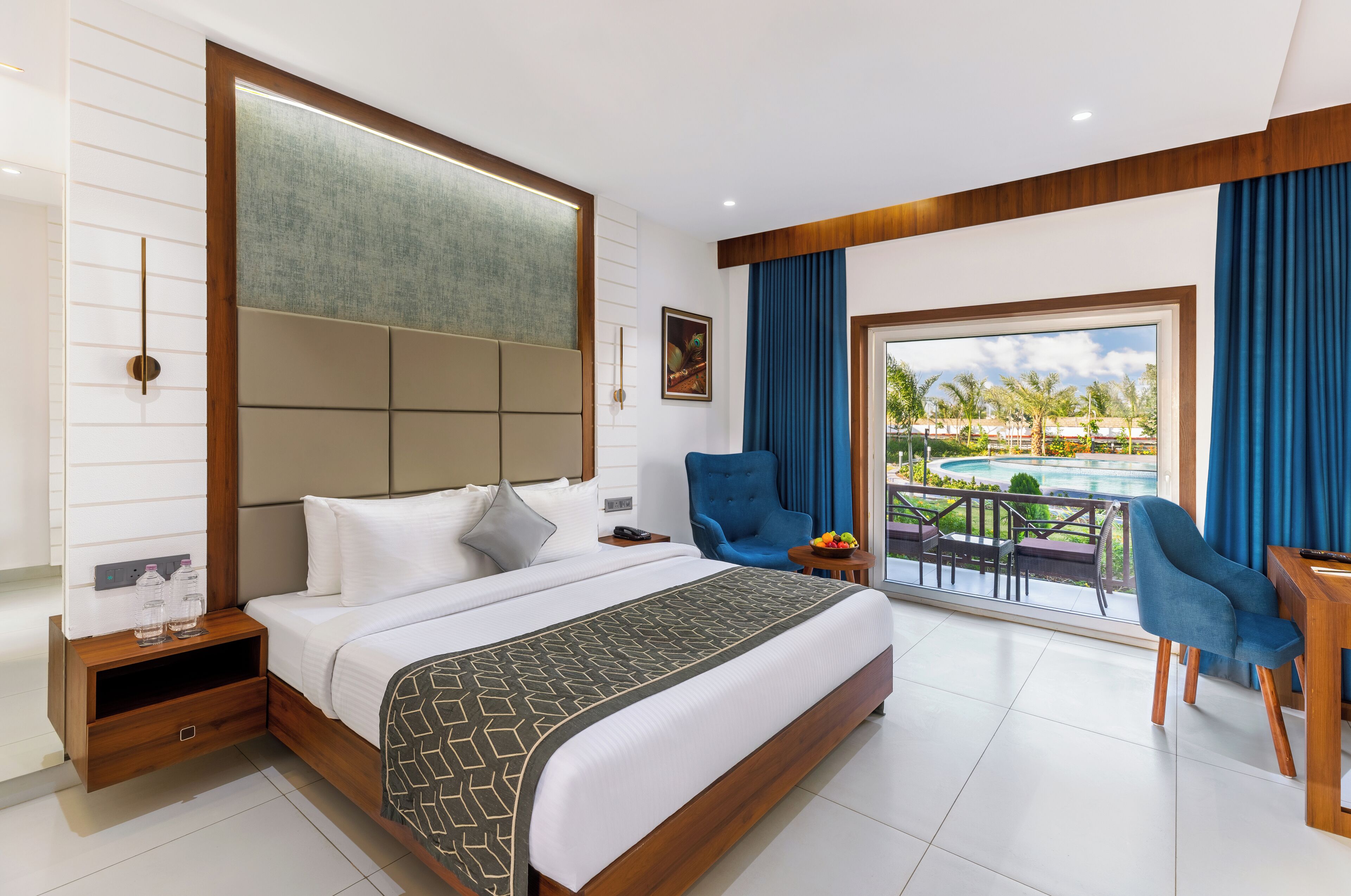 Quarto de hotel moderno com uma cama grande, vista para uma piscina e uma varanda com cortinas azuis.