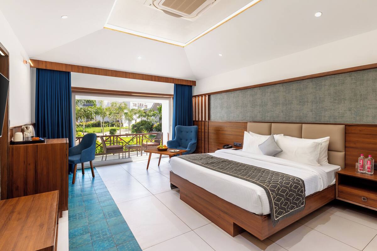 A modern hotel room with a large bed, a desk, a chair, and a balcony with a view of greenery.