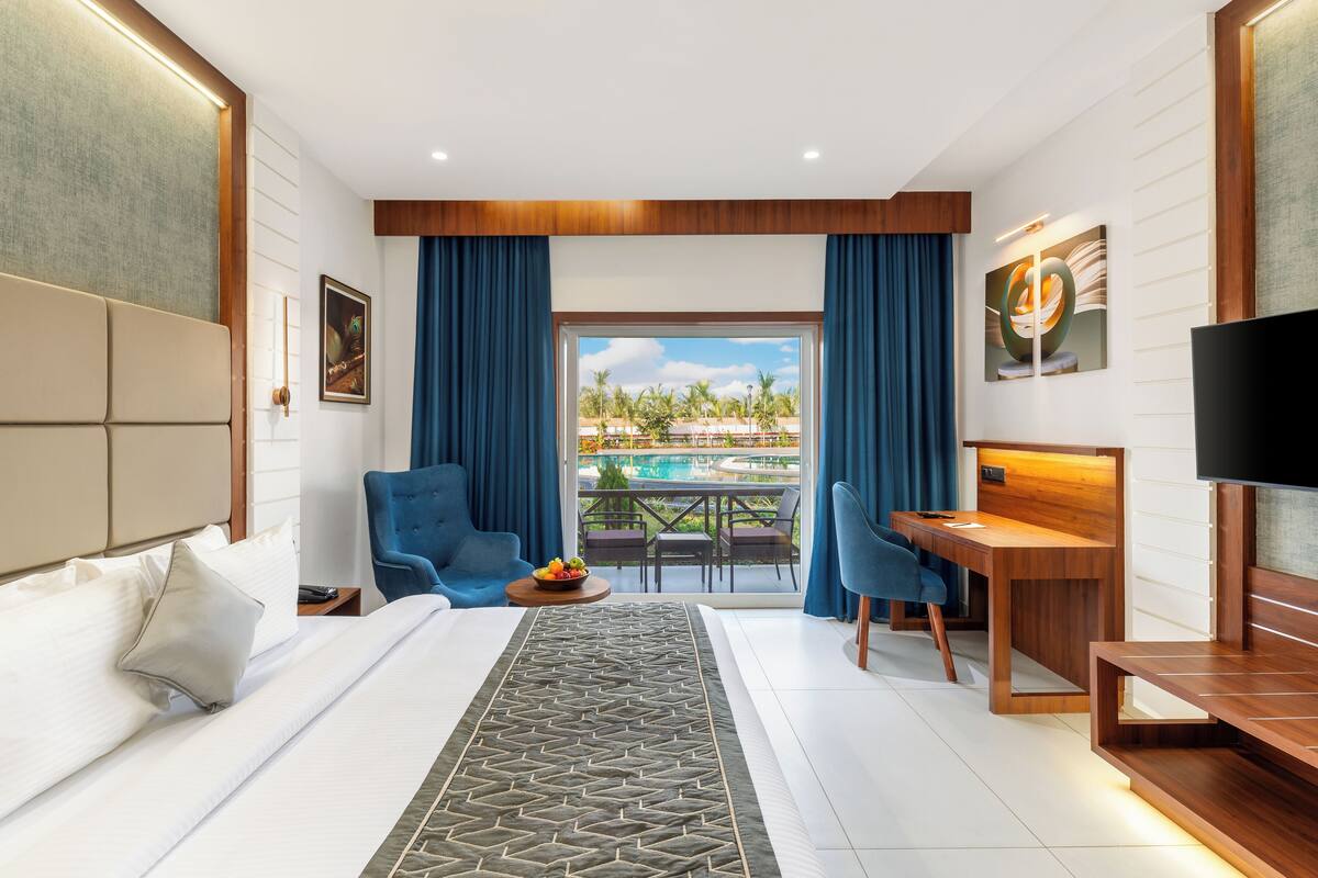 A hotel room with a bed, a desk, a TV, and a balcony with a view of palm trees.