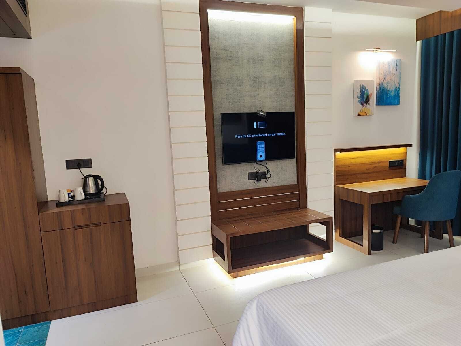 A modern hotel room with a bed, a desk, a TV mounted on a wooden panel, and a coffee maker on the side table.