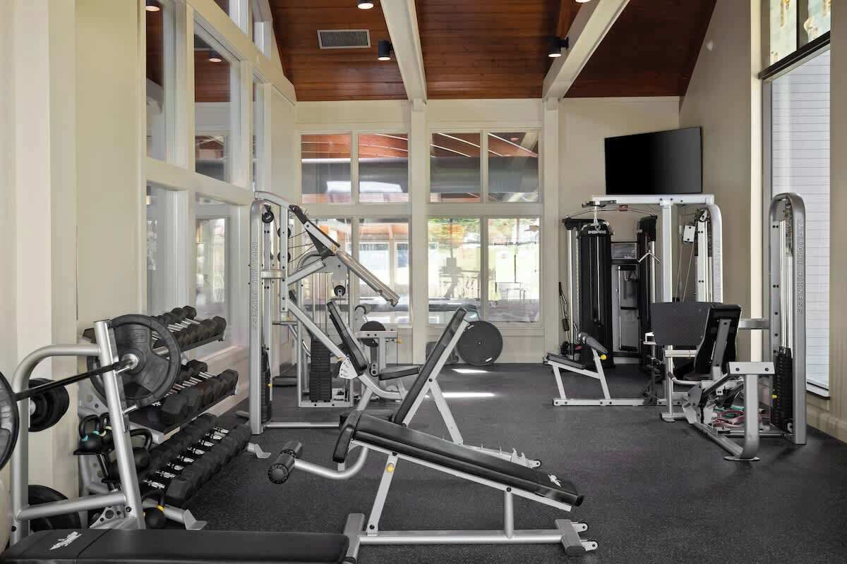 Ein Fitnessstudio mit verschiedenen Trainingsgeräten, großen Fenstern und einem Fernseher an der Wand.