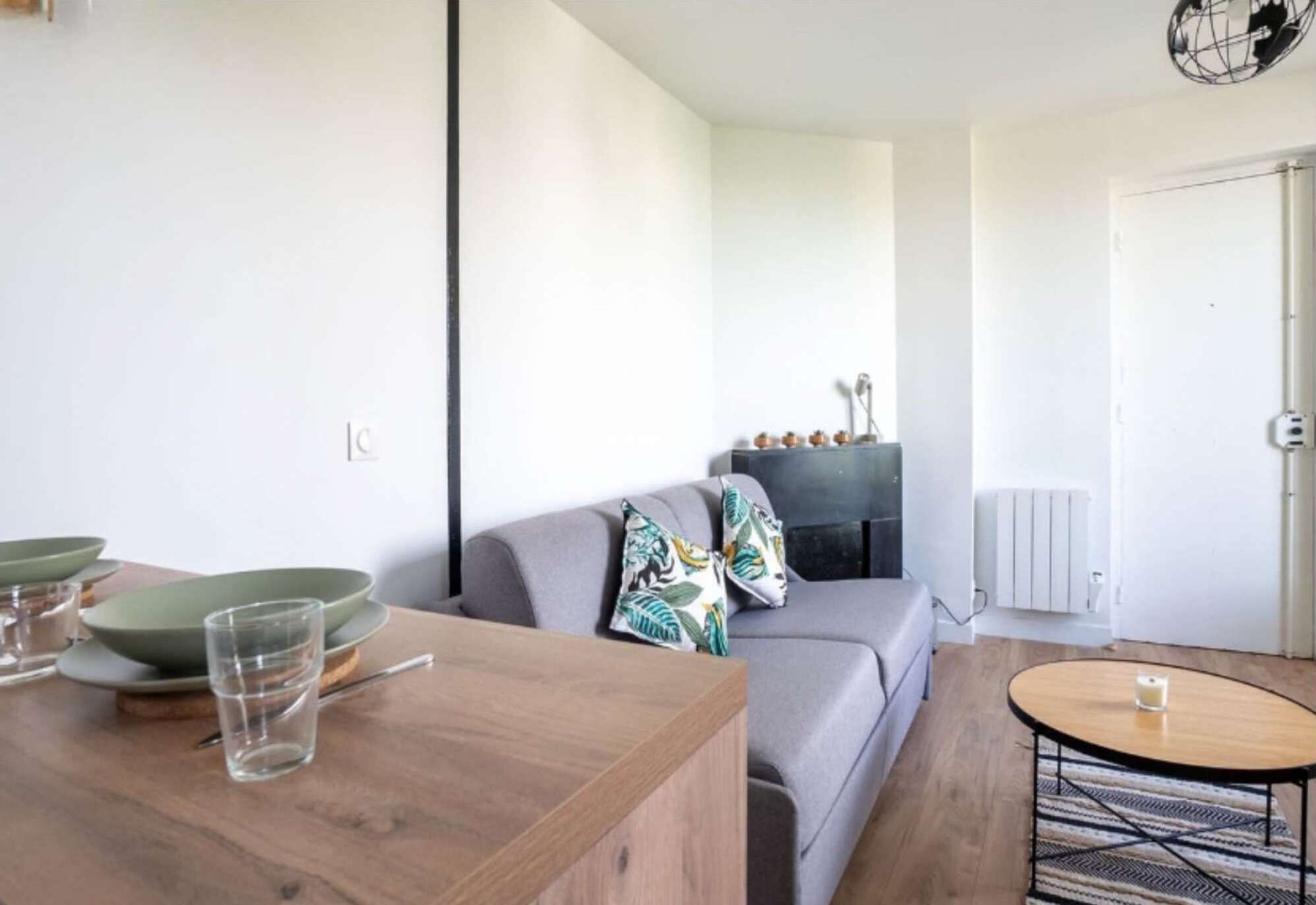 A modern living room with a wooden dining table, a grey sofa with patterned cushions, and a small round table with a candle.
