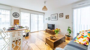 En lys stue med spiseplads, fjernsyn og sofa med et farverigt pude.