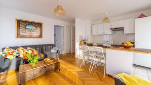 En moderne lejlighed med stue, køkken og spiseområde. Der er en sofa, et spisebord med stole og en køkkenbordplade med en skål frugt.