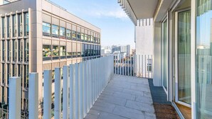 En moderne bygning med en glasfacade, et overdækket udendørs område med et metalhegn og udsigt til andre bygninger i baggrunden.