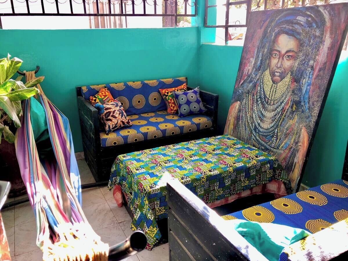 A room with a teal wall, a sofa with patterned cushions, a small table with a colorful tablecloth, and a large painting.