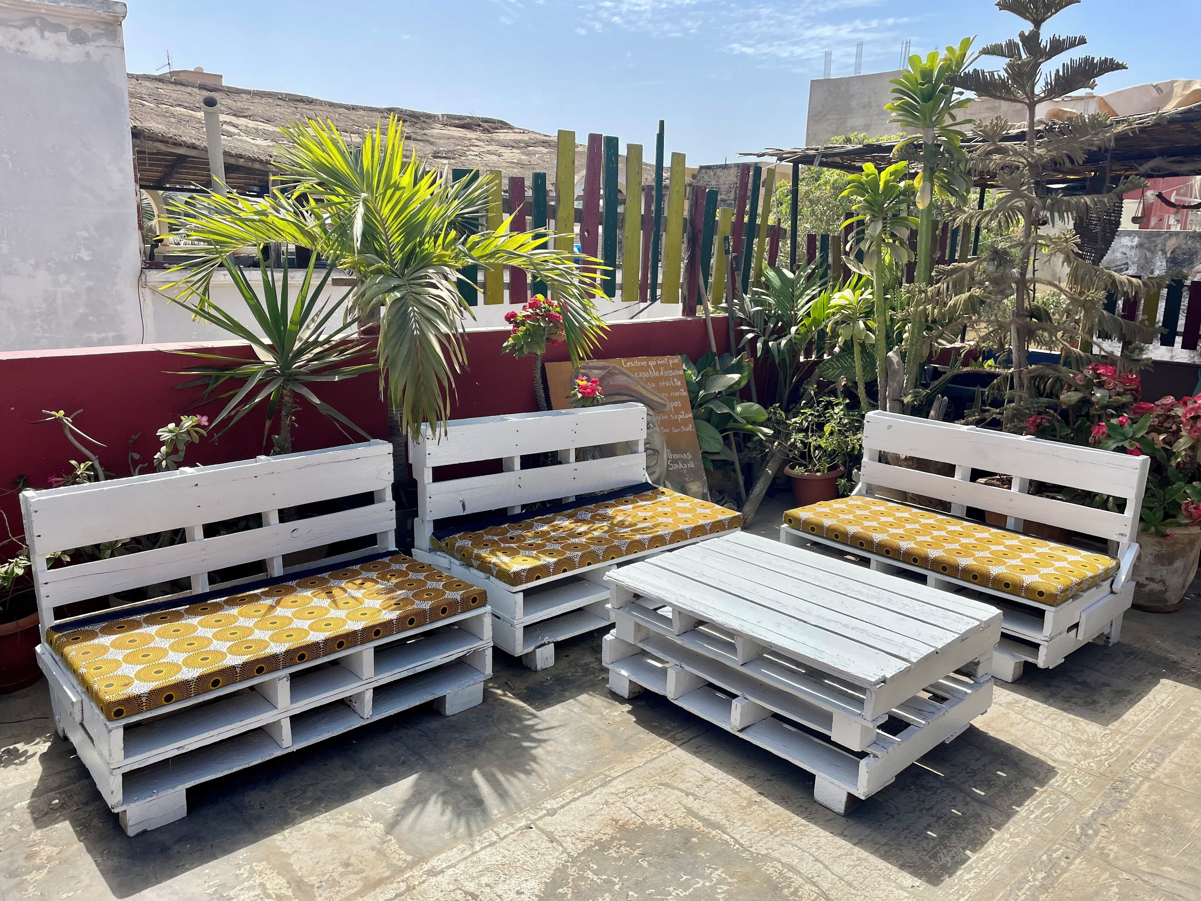 Outdoor seating with white benches and patterned cushions, surrounded by potted plants and a red wall.