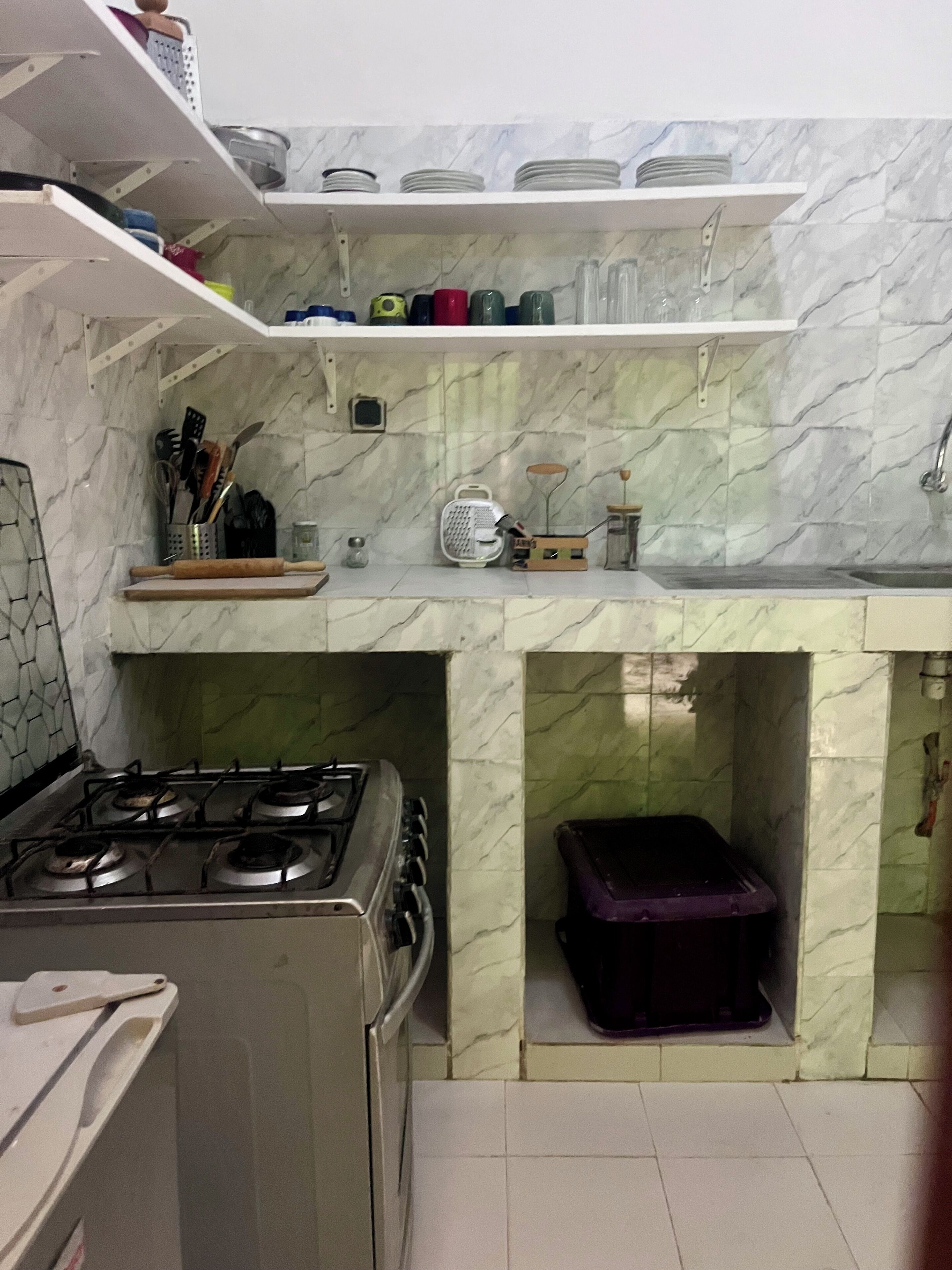 Una cocina con estufa a gas, lavamanos y salpicadero de mármol. Hay repisas sobre el lavamanos con platos y tazas.
