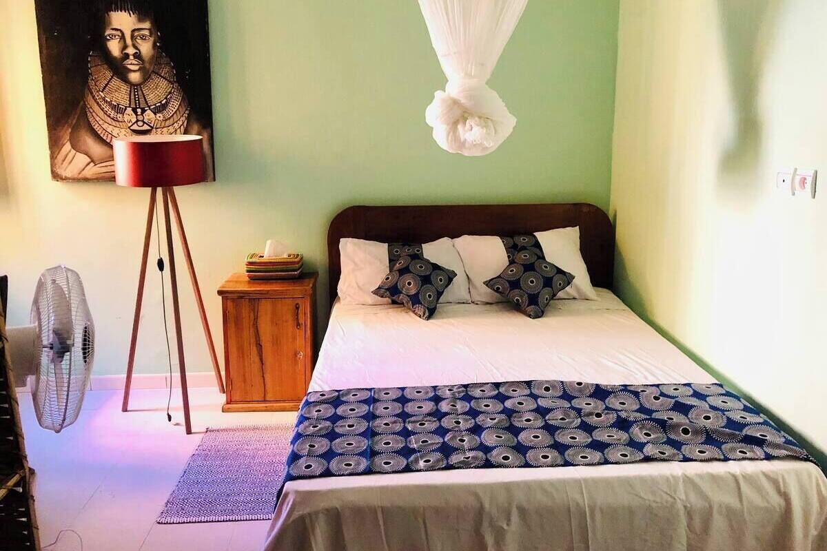 A bedroom with a bed, a bedside table, a lamp, a fan, and a painting on the wall.