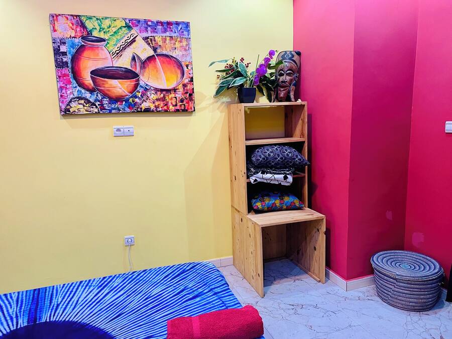 A room with a colorful painting, a wooden shelf with cushions, a potted plant, and a red wall.