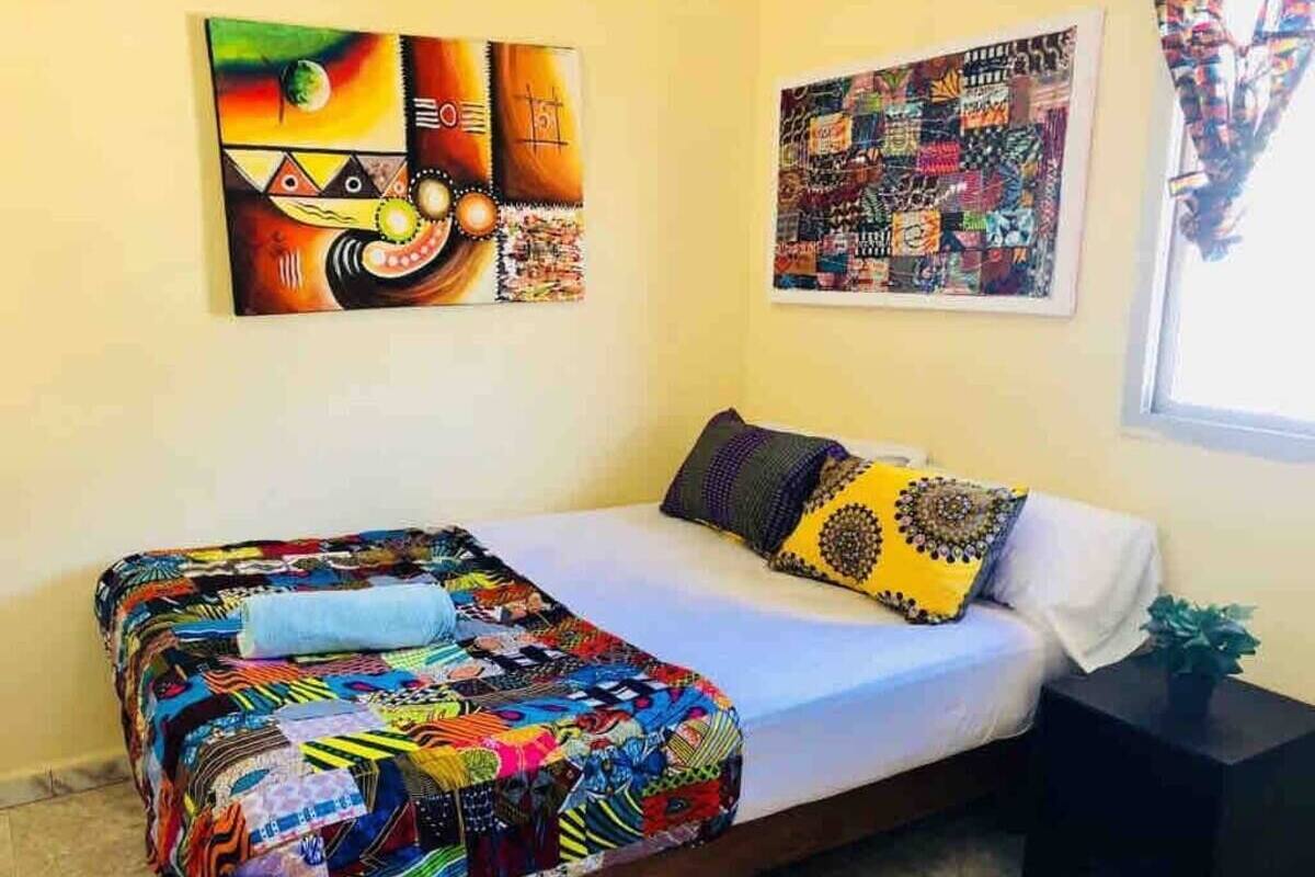 A single-bed room with a colorful quilt, a small bedside table, and two wall art pieces.