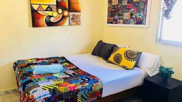 Habitación con cama individual, colcha colorida, una mesita de noche pequeña y dos obras de arte en la pared.