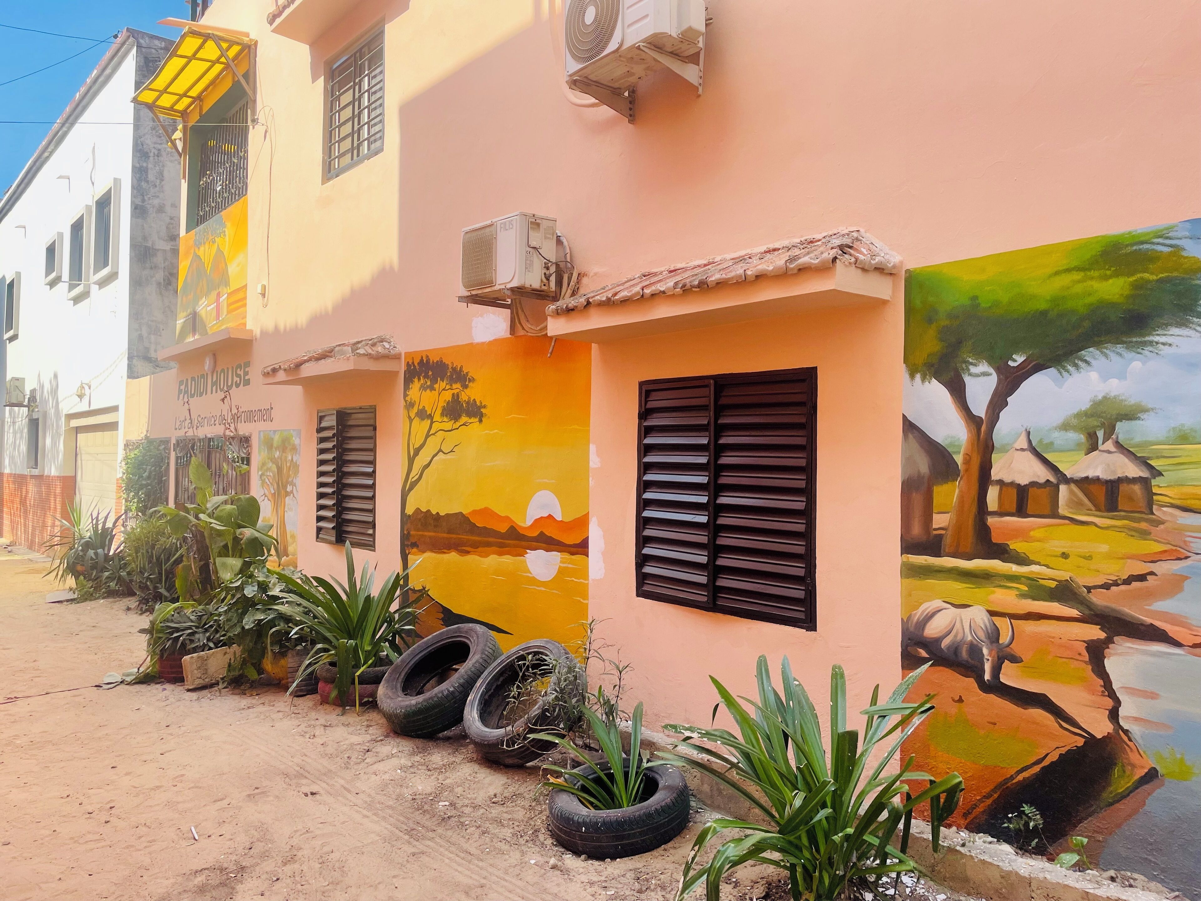 A building with a mural of a sunset and a cow, a sign reading "FADIDI HOUSE," and several potted plants and tires on the ground.