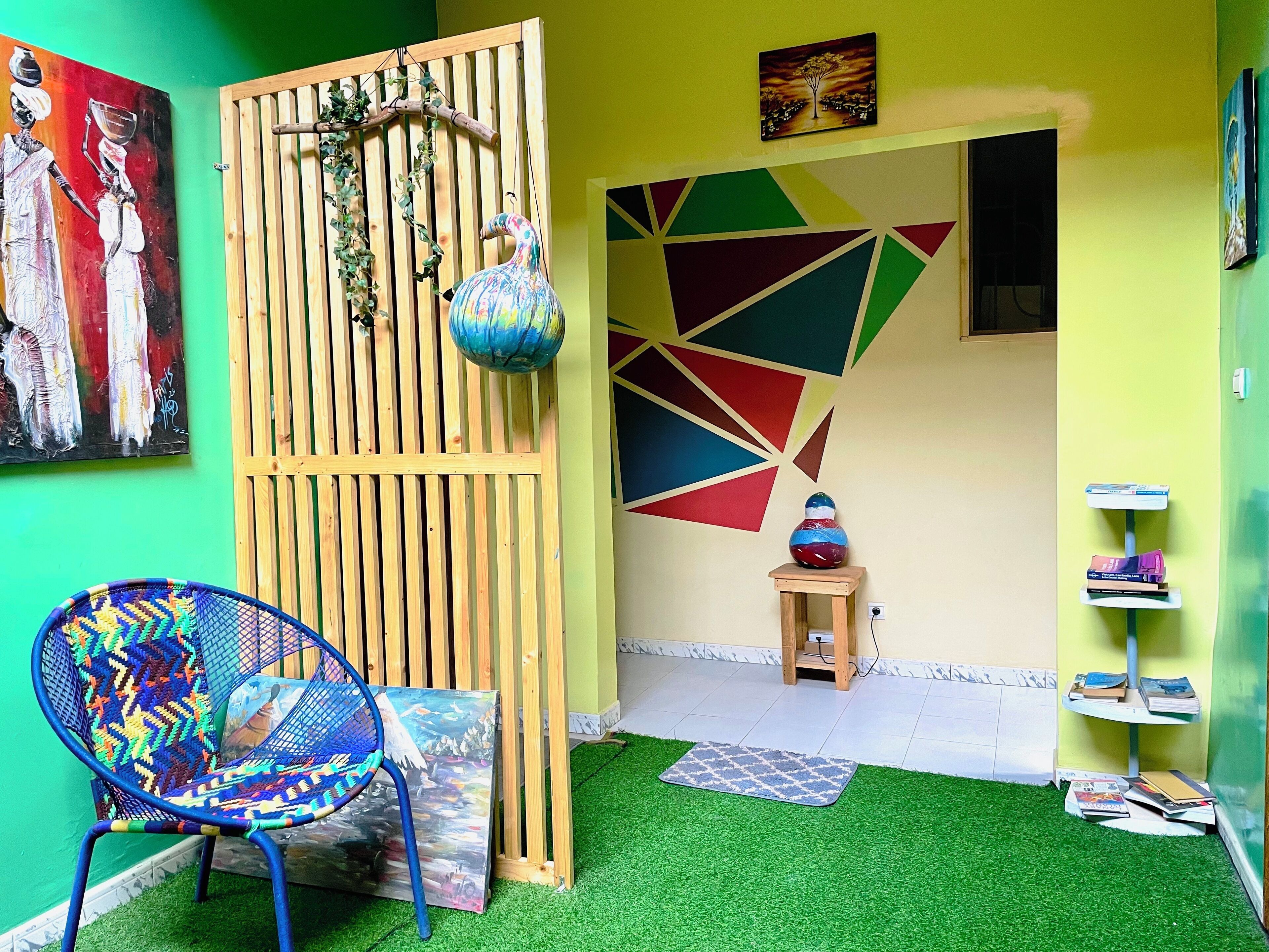A room with a green wall, a wooden partition with hanging plants, a colorful chair, a small wooden table with books, and a geometric wall design.