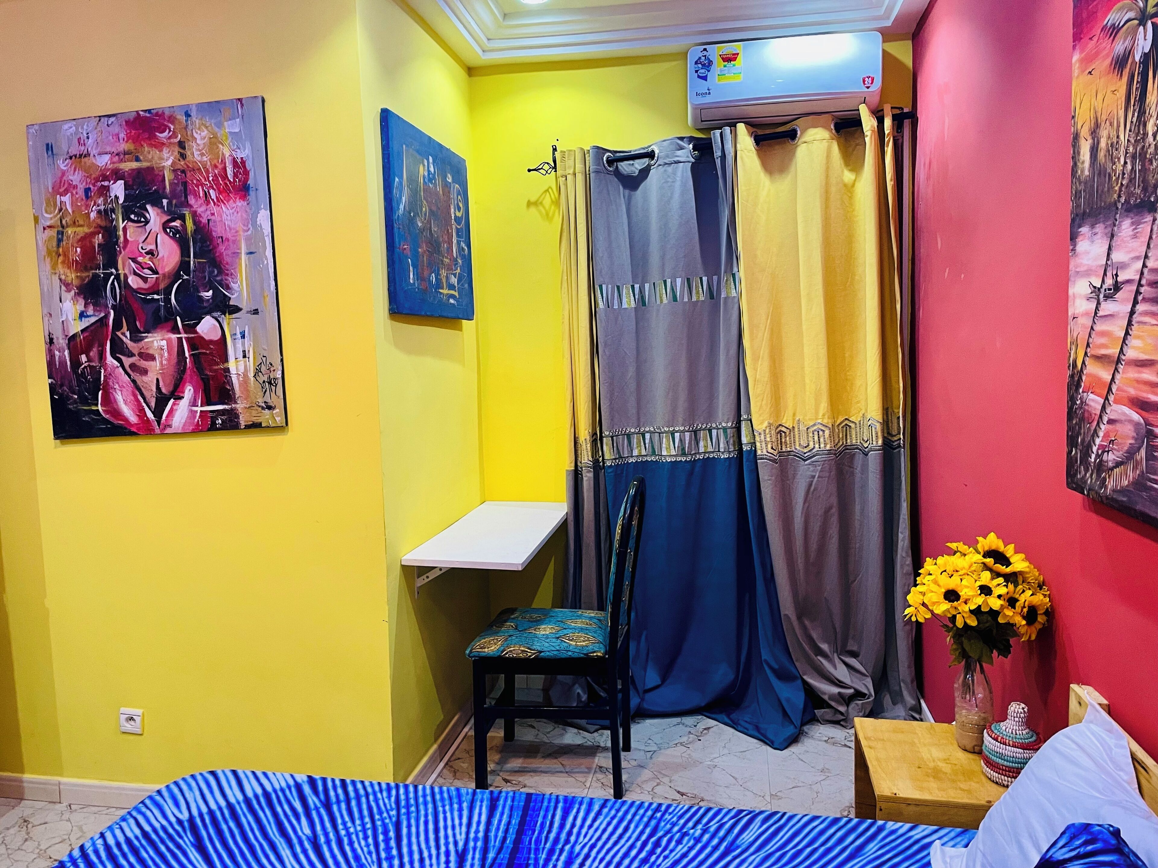 A bedroom with a bed, a chair, a small table, a wall-mounted shelf, a shower curtain, and two paintings.