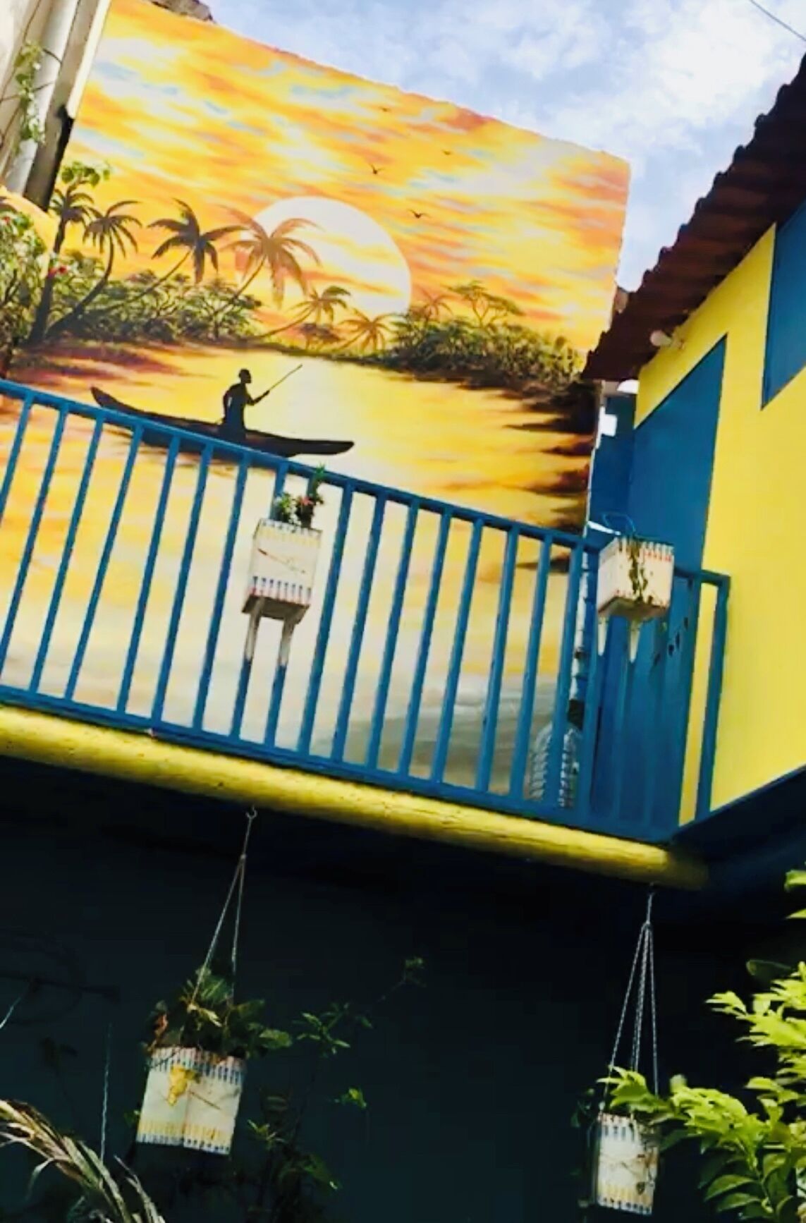 A building with a mural of a sunset over water and palm trees, a blue railing, and hanging planters.