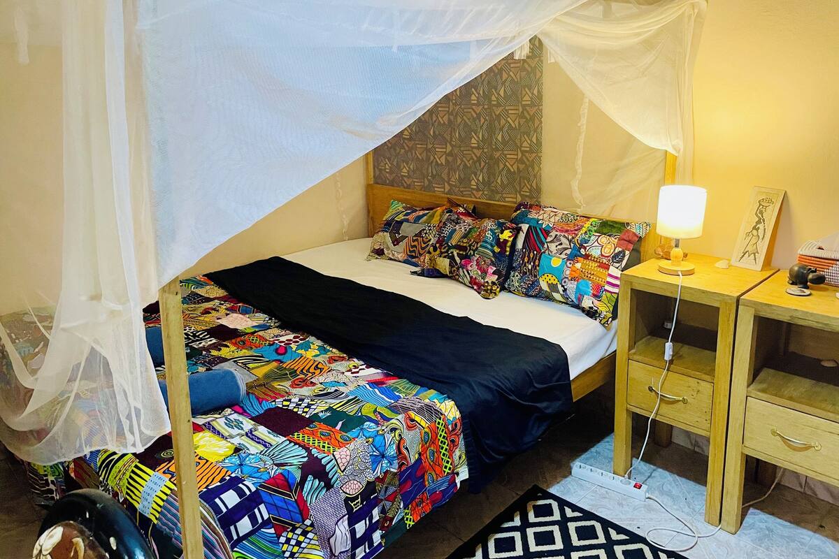 A room with a bed covered in colorful bedding, a bedside table with a lamp, and a small drawer unit.