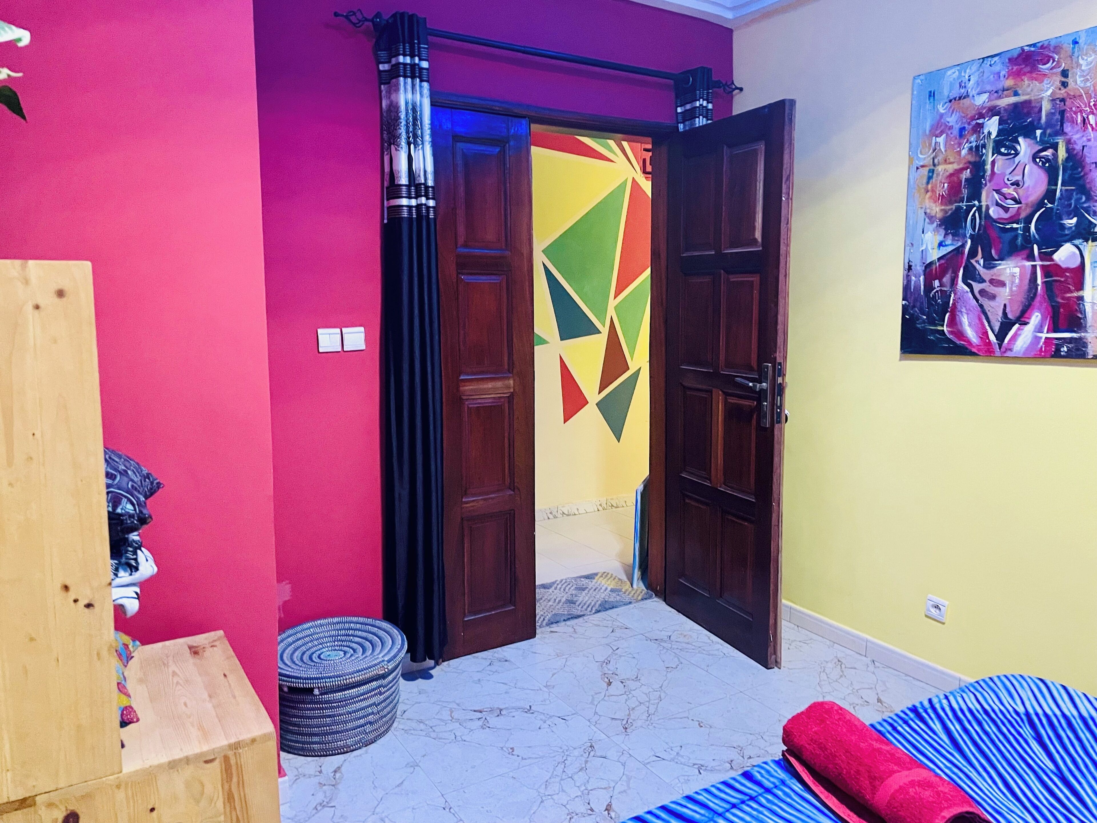 A room with a wooden wardrobe, a bed with a blue and red striped bedspread, a colorful geometric wall design, and a painting on the wall.