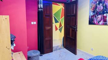 A room with a wooden wardrobe, a bed with a blue and red striped bedspread, a colorful geometric wall design, and a painting on the wall.