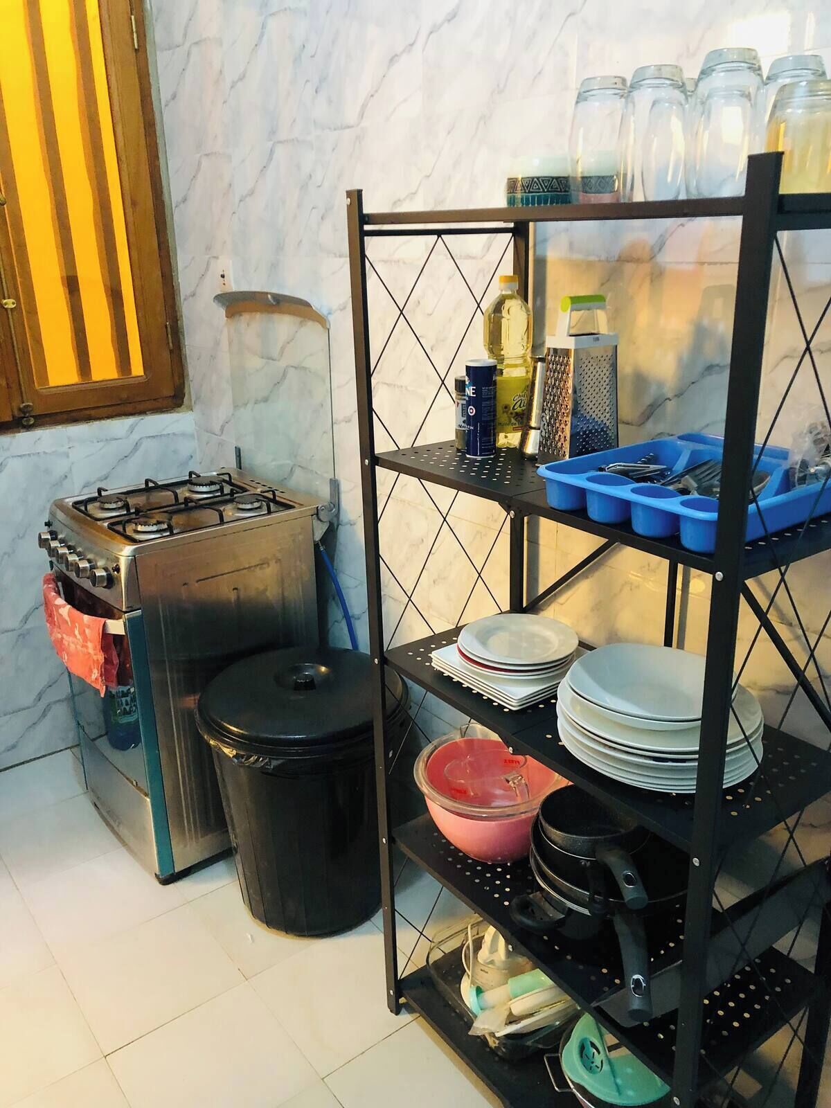 A kitchen area with a gas stove, a trash bin, a shelf with kitchen utensils, and a shelf with jars and bottles.