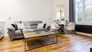 A modern living room with a grey sofa, a coffee table, and a dining area with white chairs and a table. - Peaceful Apartment - 1br/4p - Eiffel Tower (Paris)