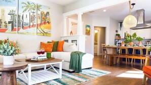 Una sala de estar moderna con un sofá blanco, cojines coloridos, una mesa de centro y un comedor con sillas y una mesa de madera.
