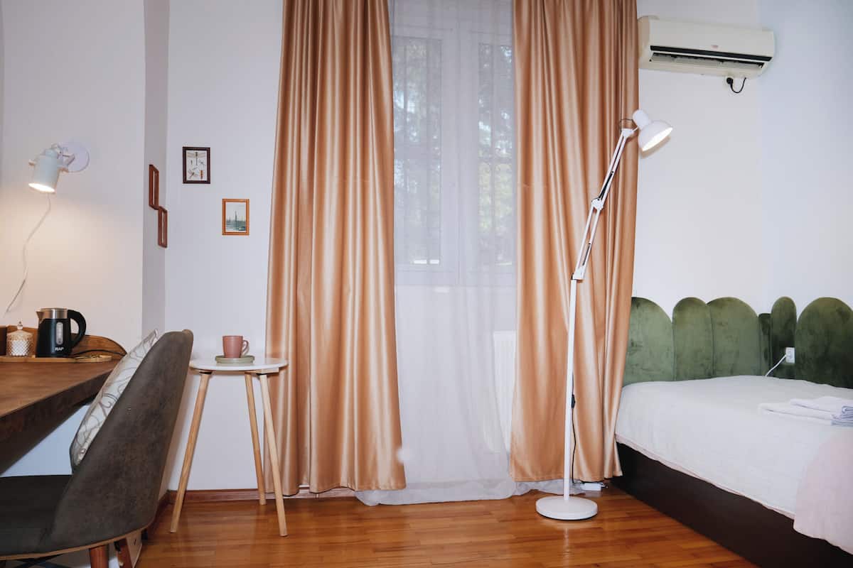 Habitación de hotel con piso de madera, una cama con cabecera verde, un escritorio con una silla, una mesita con una taza y una lámpara.